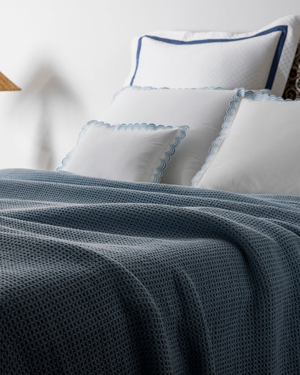 Slate blue waffle-weave Chatham blanket draped over white sheets with blue scalloped trim.