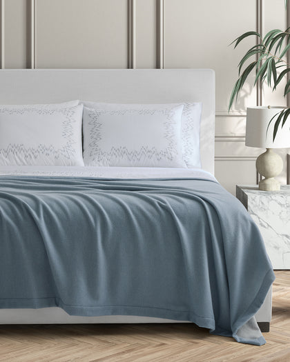 Slate blue Cosmo blanket draped over a bed with zigzag embroidered white shams.
