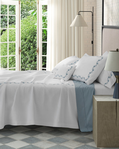 White Daphne Matelasse quilted bedding with floral embroidery, layered over blue sheets.