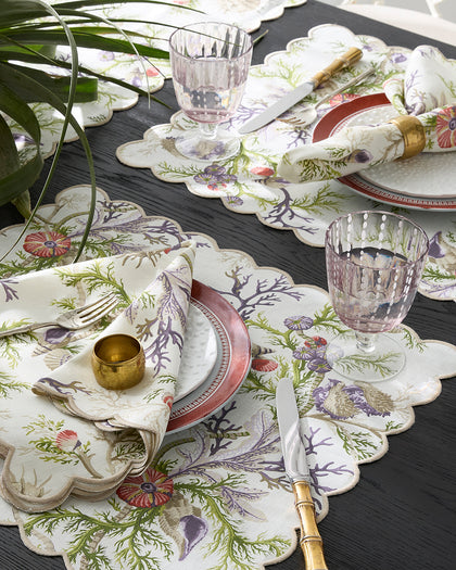 Del Tesoro coral and shell print scalloped placemats and napkins, set on a table with gold flatware.