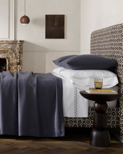Charcoal Dream Modal sheet draped over white embroidered sheets, styled beside a fireplace.