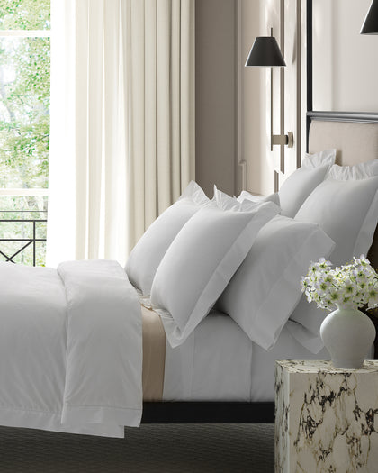 Ivory Gatsby Hemstitch bedding with ruffled shams, styled beside white flowers.