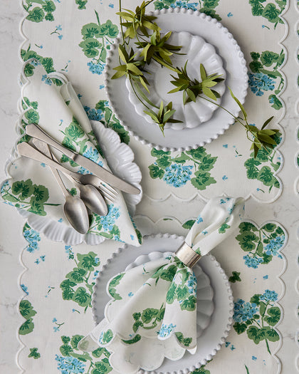 Blue and green Geranium print scalloped placemats and napkins, set with white scalloped plates from above.