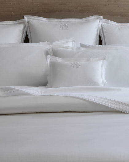 White Grace monogrammed pillows with woven lace trim, styled against a headboard.