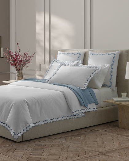 White India scalloped sheets with blue trim, layered with a blue throw beside cherry blossom branches.