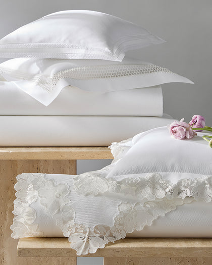 White Lace Pique bedding with floral lace trim, stacked on a bench beside ranunculus.