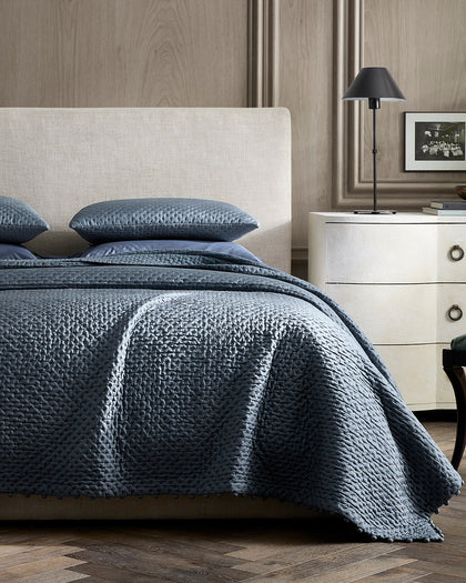 Slate blue Margaret textured quilted bedding with a pompom trim, styled beside a nightstand.