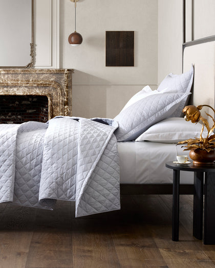 Pale gray diamond-quilted Milano Quilt bedding styled beside a fireplace.