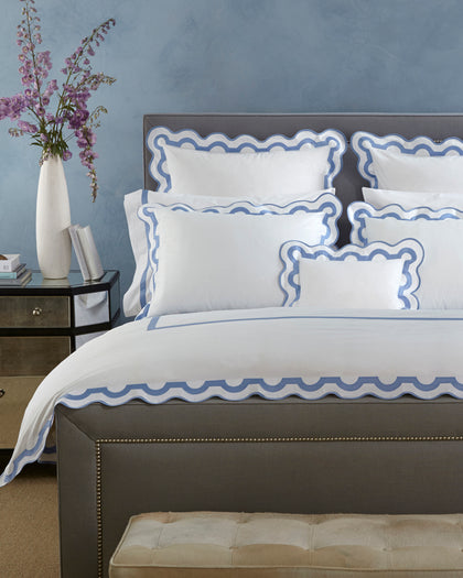 White Mirasol bedding with a blue scalloped multicolor trim, styled on a bed.