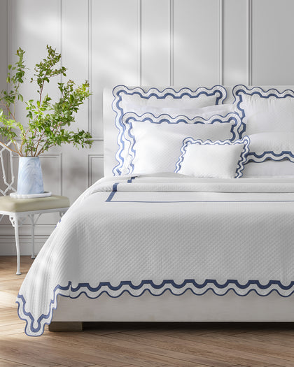 White Mirasol Matelasse quilted bedding with a navy scalloped trim, styled beside a bench.