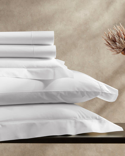 White Positano Hemstitch sheets and pillows stacked beside a dried protea flower.