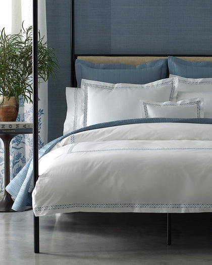 White Prado sheets with zigzag embroidered trim, layered with blue quilted pillows on a bed.