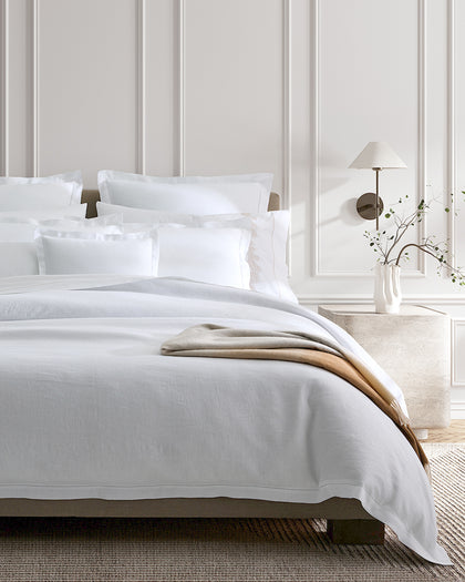 White linen Roman Hemstitch bedding with a tan throw draped over the corner.