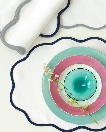 White Scallop Edge placemats with navy and gray trim, set with teal and pink stacked plates from above.