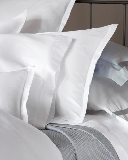 White Talita Satin Stitch pillows with a satin stitch border, layered with silver-trimmed shams on a bed.