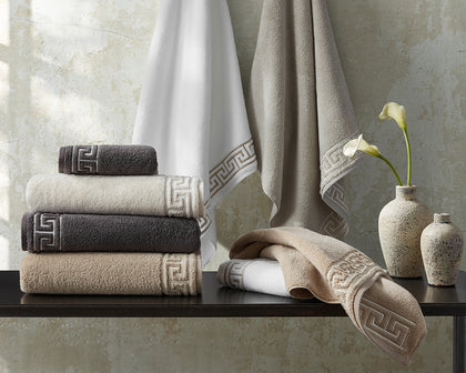 Adelphi towels with greek-key trim in white, tan, and charcoal, hung and stacked beside calla lilies.