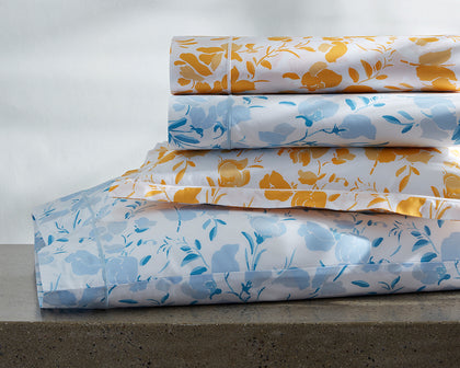 Alexandra floral print sheets in gold and blue, stacked on a stone ledge.