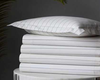 White Amalfi pinstripe sheets stacked on a marble table beside a palm frond.