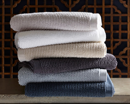 Ribbed Aman towels stacked in white, tan, gray, and navy, in front of a carved wood screen.