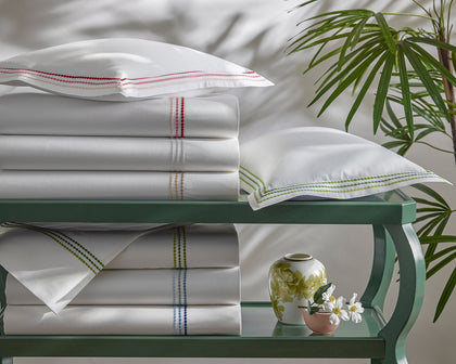 White Amara Triple Pearl sheets with triple-dot embroidered trim in assorted colors, stacked on a green table.