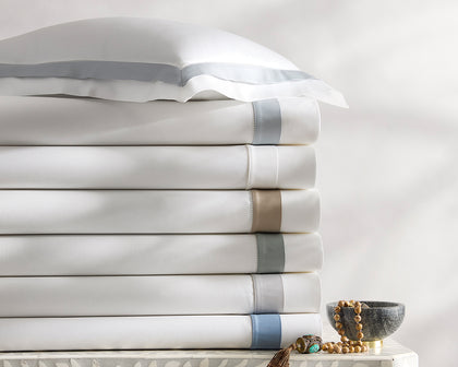 White Ambrose sheets with satin bordered trim in blue and tan, stacked on an inlaid dresser.
