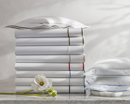 White Ansonia sheets with single-stripe trim in assorted colors, stacked beside a white poppy.