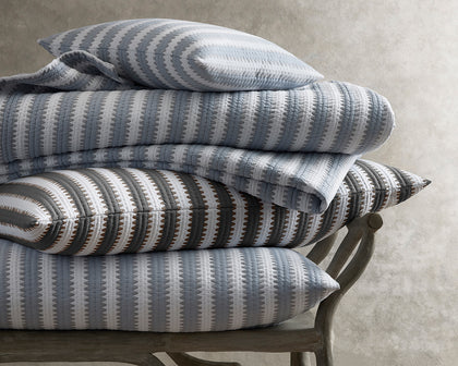 Apollo Stripe Matelasse quilted bedding in blue and gray, stacked on a wooden bench.