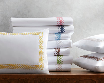 White Astor Braid sheets and pillow with loop-embroidered trim in gold and assorted colors, stacked on a wooden table.