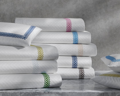 White Astor Braid quilted bedding with loop-embroidered trim in a rainbow of colors, stacked on a marble table.