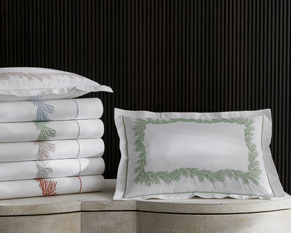 White Atoll sheets with coral-branch embroidered trim in assorted colors, stacked beside a green-embroidered sham.