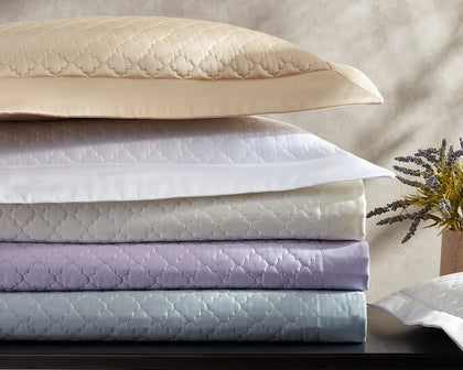 Ava quatrefoil-quilted bedding stacked in gold, white, lavender, and blue, beside a sprig of lavender.