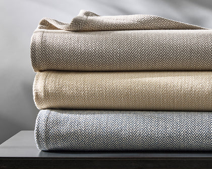 Avalon herringbone-woven blankets folded in gray, tan, and blue, stacked on a dark table.