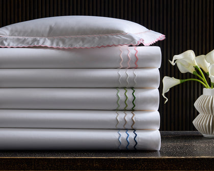 White Axel sheets with wavy embroidered trim in assorted colors, stacked beside calla lilies.