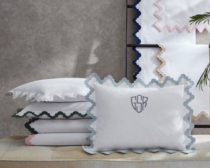 White Aziza pillow with blue wavy trim and a monogram, stacked with sheets in assorted colors.