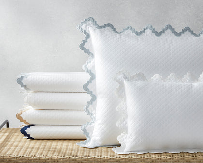 White Aziza Matelasse quilted bedding with gray wavy trim, stacked on a woven bench.