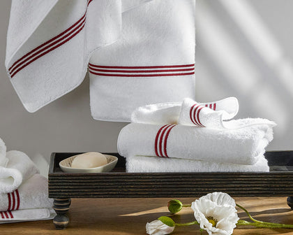 White Bel Tempo towels with red triple-stripe trim, hung and stacked beside a poppy flower.