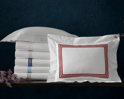 Stacked white Bel Tempo sheets with colorful striped trim beside a red-bordered euro sham.