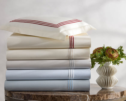 Stacked Bel Tempo Nocturne sheets with striped embroidered trim in ivory and pale blue, beside a vase of flowers.