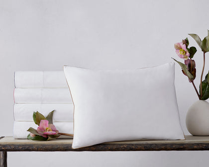 Stacked white Bryant sheets with colored piped trim, beside a tan-piped pillow and a pink flower.