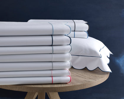 White Butterfield scalloped sheets stacked with colored trim, beside a navy-trimmed sham.