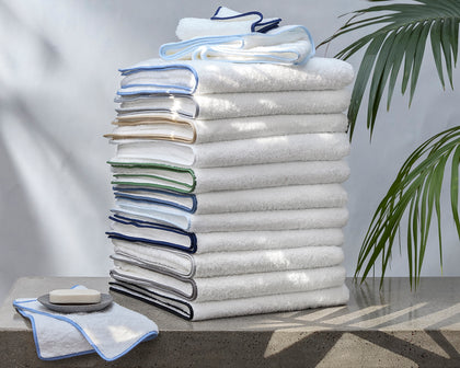 Tall stack of white Cairo towels with piped trim in a rainbow of colors, beside soap and a palm plant.