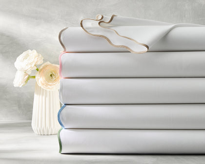 White Camilla Pique sheets stacked with colored edge trim, beside a vase of white ranunculus.