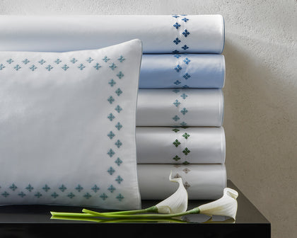 White Casablanca sheets with diamond cross-stitch trim in assorted colors, stacked beside calla lilies.