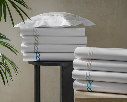 White Classic Chain sheets stacked with chain-link embroidered trim in assorted colors, near a plant.