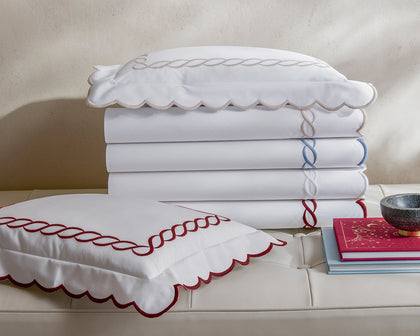 White Classic Chain Scallop sheets with scalloped chain-embroidered trim in tan, blue, and red, stacked on a tufted bench.