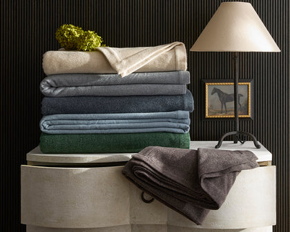 Cosmo blankets folded in tan, gray, navy, and green, stacked on a chest beside a horse painting.