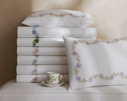 White Daphne sheets stacked with embroidered floral trim in assorted colors, beside a teacup.