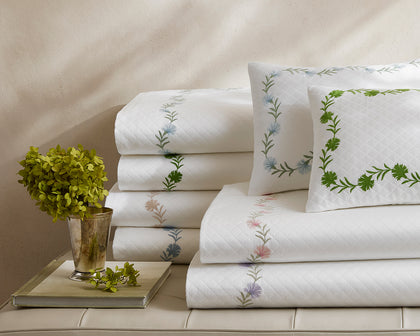 White Daphne Matelasse quilted sheets with floral embroidery, stacked on a bench beside a plant.