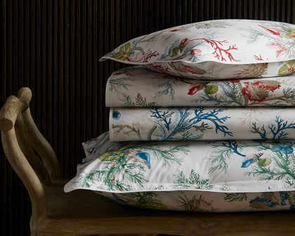 Del Tesoro coral and shell print sheets in red and blue, stacked on a wooden bench.