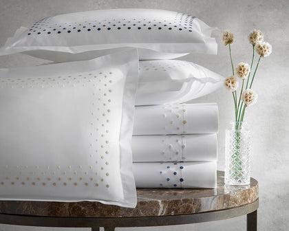 White Dorothy sheets with polka-dot embroidered borders, stacked beside a vase of dried flowers.